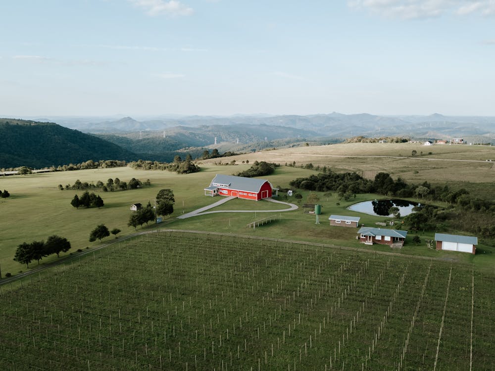 An aerial view of Tyn-yr-Onnen Farm Campsite in Snowdonia National Park, showing a red barn, a pond, and several small buildings surrounded by vast green fields. The scene is framed by rolling hills and distant mountains under a clear blue sky.