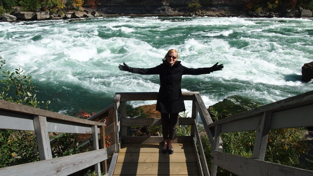 A woman in a black coat stands with her arms outstretched on a wooden platform overlooking the turbulent, foamy rapids of a wide river. The scene is framed by the steep steps of a wooden staircase leading down to the platform.