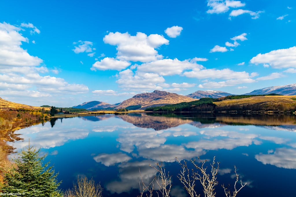A tranquil lake reflects a vibrant blue sky dotted with fluffy white clouds. Surrounding the lake, rolling hills and distant mountains create a picturesque landscape, with green trees and brush on the shore adding to the natural beauty. Not far off, River Spean meanders gracefully through this serene scene.