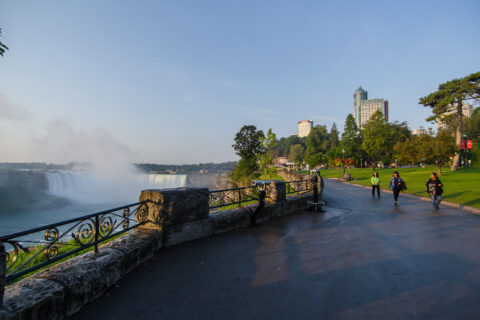 People stroll along a scenic path by a waterfall in Queen Victoria Park, Ontario, Canada, surrounded by lush greenery.